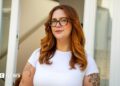Jess King, a woman with long ginger hair and glasses, stands in front of a doorway. She is wearing a short-sleeved white top and has visible black tattoos at the top of her arms.