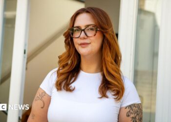 Jess King, a woman with long ginger hair and glasses, stands in front of a doorway. She is wearing a short-sleeved white top and has visible black tattoos at the top of her arms.