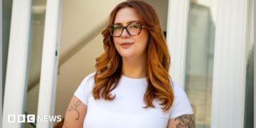 Jess King, a woman with long ginger hair and glasses, stands in front of a doorway. She is wearing a short-sleeved white top and has visible black tattoos at the top of her arms.