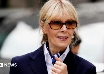 E Jean Carroll with a navy blazer, white shirt and blue patterned scarf. She is wearing large sunglasses and looking straight ahead.