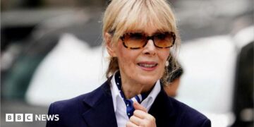E Jean Carroll with a navy blazer, white shirt and blue patterned scarf. She is wearing large sunglasses and looking straight ahead.