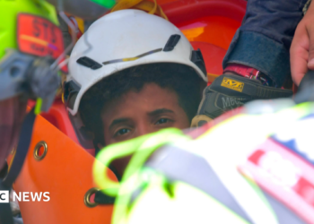 the picture shows half of the boys face - the rest is obscured by rescue workers. He's wearing a white hard hat and has dark curly hair.