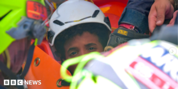 the picture shows half of the boys face - the rest is obscured by rescue workers. He's wearing a white hard hat and has dark curly hair.