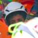 the picture shows half of the boys face - the rest is obscured by rescue workers. He's wearing a white hard hat and has dark curly hair.