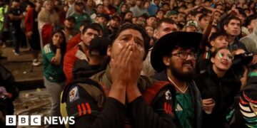 A man is holding his hands over his mouth with numerous of Mexican fans behind him.