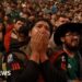 A man is holding his hands over his mouth with numerous of Mexican fans behind him.