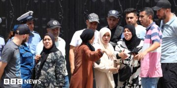 Police stand guard at a black gates of an orphanage in Algiers as a crowd of men and women gesticulate outside