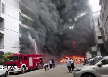 Plumes of black smoke and flames can be seen as a fire truck sprays a burning building with water