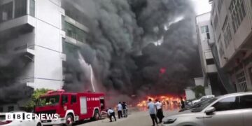 Plumes of black smoke and flames can be seen as a fire truck sprays a burning building with water