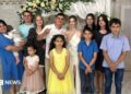 Twelve family members smile at the camera while what appears to be a bride and groom hold up their traditional wedding ring fingers