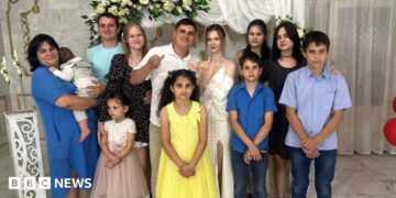 Twelve family members smile at the camera while what appears to be a bride and groom hold up their traditional wedding ring fingers