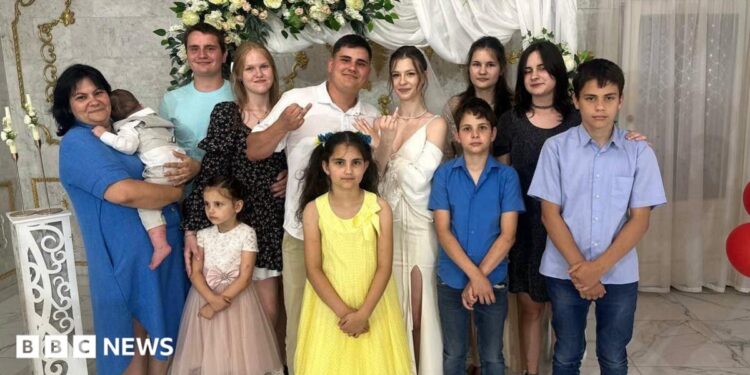 Twelve family members smile at the camera while what appears to be a bride and groom hold up their traditional wedding ring fingers