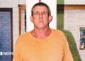 A tall man in a yellow t-shirt and blue short stands against a measuring tape in an office