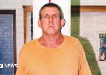 A tall man in a yellow t-shirt and blue short stands against a measuring tape in an office