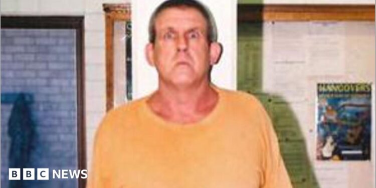 A tall man in a yellow t-shirt and blue short stands against a measuring tape in an office