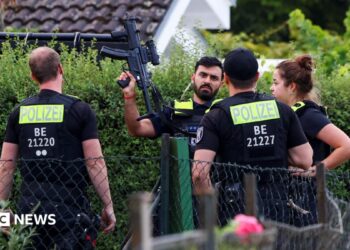 Four police officers are pictured, with one officers holding a gun up