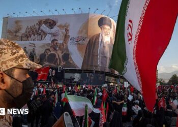 an Iranian security officer amid crowds in front of a billboard showing the late supreme leader at his funeral
