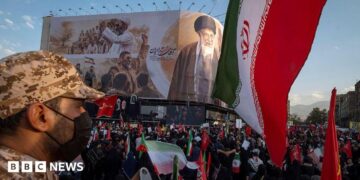 an Iranian security officer amid crowds in front of a billboard showing the late supreme leader at his funeral
