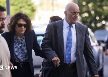 A balding man in a grey suit holds the hand of a woman in a navy blue suit who is wearing sunglasses as both are walking