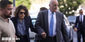 A balding man in a grey suit holds the hand of a woman in a navy blue suit who is wearing sunglasses as both are walking