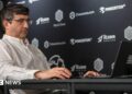 Vladimir Kramnik at a chess tournament in London in August 2024. He sits facing his opponent and is wearing a white shirt and glasses.