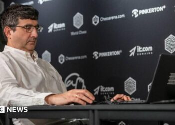 Vladimir Kramnik at a chess tournament in London in August 2024. He sits facing his opponent and is wearing a white shirt and glasses.