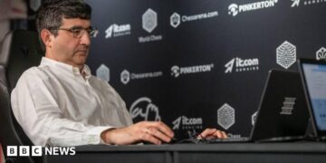 Vladimir Kramnik at a chess tournament in London in August 2024. He sits facing his opponent and is wearing a white shirt and glasses.