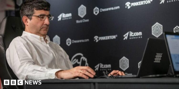 Vladimir Kramnik at a chess tournament in London in August 2024. He sits facing his opponent and is wearing a white shirt and glasses.