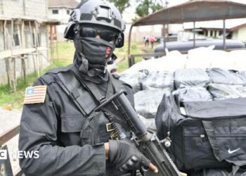 An armed Liberian police officer stands guard beside a truck carrying bags and packages