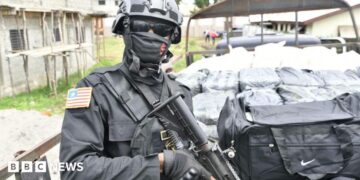 An armed Liberian police officer stands guard beside a truck carrying bags and packages
