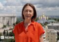 Sarah Rainsford wears a short-sleeved bright orange shirt. She reports in front of several buildings in Crimea