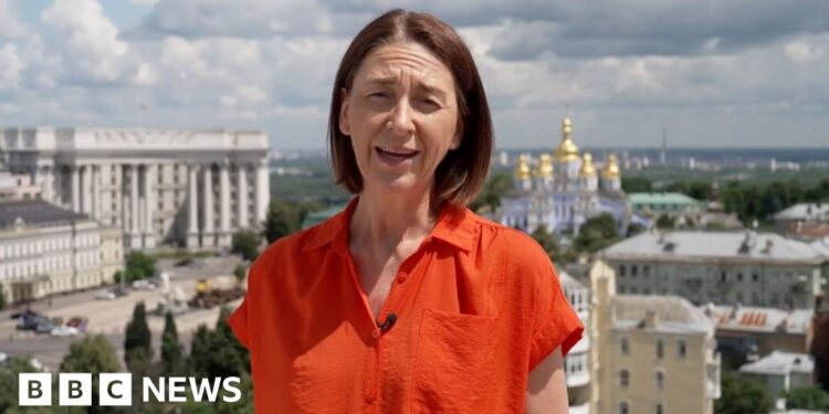 Sarah Rainsford wears a short-sleeved bright orange shirt. She reports in front of several buildings in Crimea
