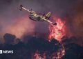 A small yellow airplane flies over red flames rising in smoke above dark trees.