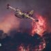 A small yellow airplane flies over red flames rising in smoke above dark trees.