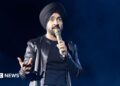 Diljit Dosanjh kicks off his Aura World Tour at BC Place on April 23, 2026, in Vancouver, British Columbia, Canada. He is wearing a black jacket over a black t-shirt and sporting a black turban, as he speaks on a microphone from the stage.