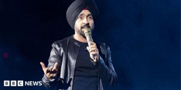 Diljit Dosanjh kicks off his Aura World Tour at BC Place on April 23, 2026, in Vancouver, British Columbia, Canada. He is wearing a black jacket over a black t-shirt and sporting a black turban, as he speaks on a microphone from the stage.