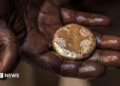 A close-up of a mud-stained hand holding a roughly circular gold nugget