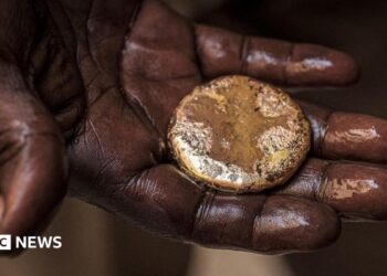 A close-up of a mud-stained hand holding a roughly circular gold nugget