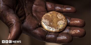 A close-up of a mud-stained hand holding a roughly circular gold nugget