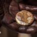 A close-up of a mud-stained hand holding a roughly circular gold nugget
