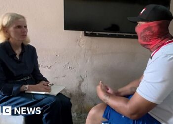 Orla Guerin sitting with a man in a white t-shirt and blue shorts, who is wearing a black baseball cap and covering his face with a red scarf.