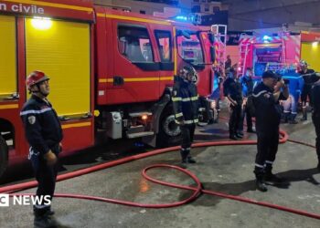 Firefighters in blue uniforms stand in front of fire trucks