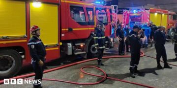 Firefighters in blue uniforms stand in front of fire trucks