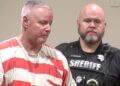 Colin Gray, in a white and orange stripped prison jumpsuit, with short grey cropped hair, is seen in profile. A sheriff's deputy in his black uniform is behind him