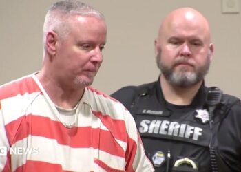 Colin Gray, in a white and orange stripped prison jumpsuit, with short grey cropped hair, is seen in profile. A sheriff's deputy in his black uniform is behind him