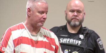 Colin Gray, in a white and orange stripped prison jumpsuit, with short grey cropped hair, is seen in profile. A sheriff's deputy in his black uniform is behind him