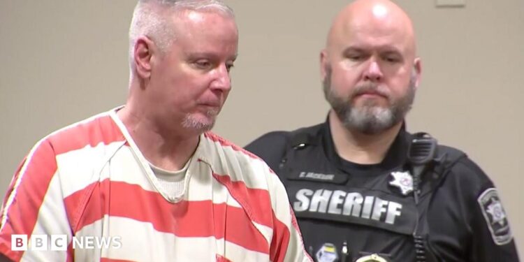 Colin Gray, in a white and orange stripped prison jumpsuit, with short grey cropped hair, is seen in profile. A sheriff's deputy in his black uniform is behind him