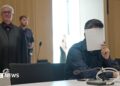 A man holds a sheet of paper over his face, wearing a black shirt in court