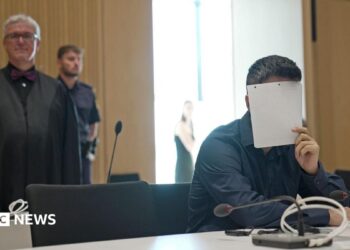 A man holds a sheet of paper over his face, wearing a black shirt in court