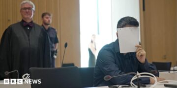 A man holds a sheet of paper over his face, wearing a black shirt in court